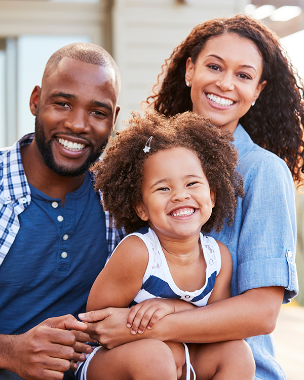 A happy family of three smiling at the camera. The father, wearing a blue plaid shirt, and the mother, in a blue dress, stand behind their young daughter, who is wearing a striped dress and sitting on someone's knee. They're outdoors in a sunny setting.