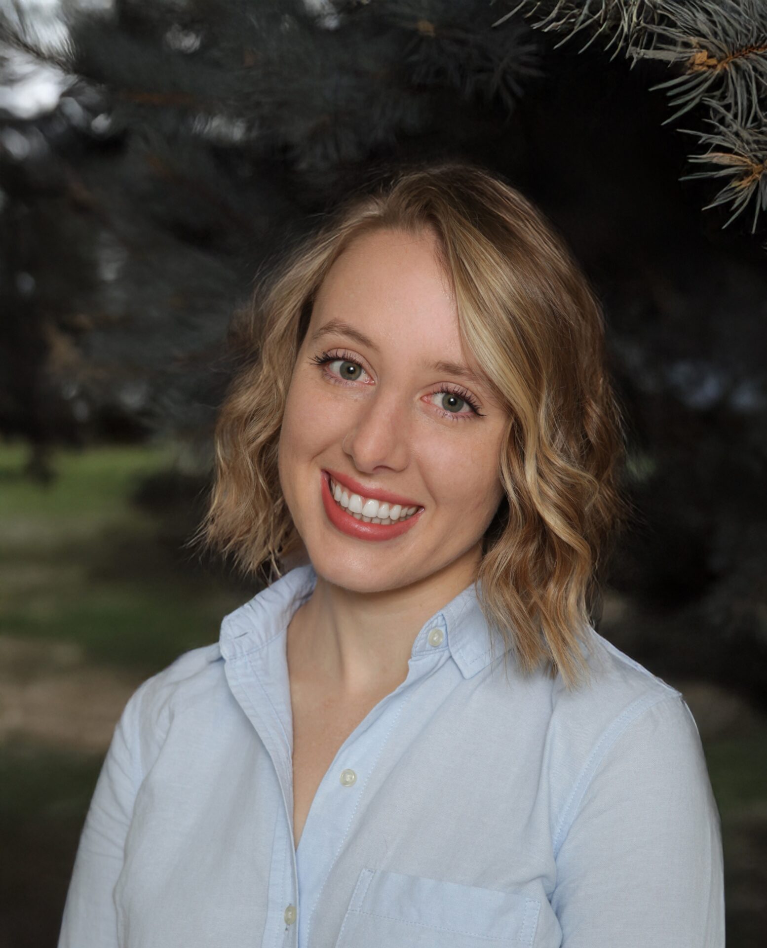 A woman with wavy blonde hair and a light blue button-up shirt smiles while standing outdoors in front of dark green trees.