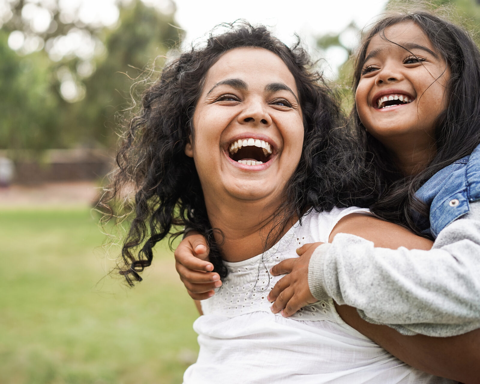 A woman and a young girl with long dark hair laugh joyfully outdoors, with the girl hugging the woman from behind. They are surrounded by greenery, and the background is blurred.