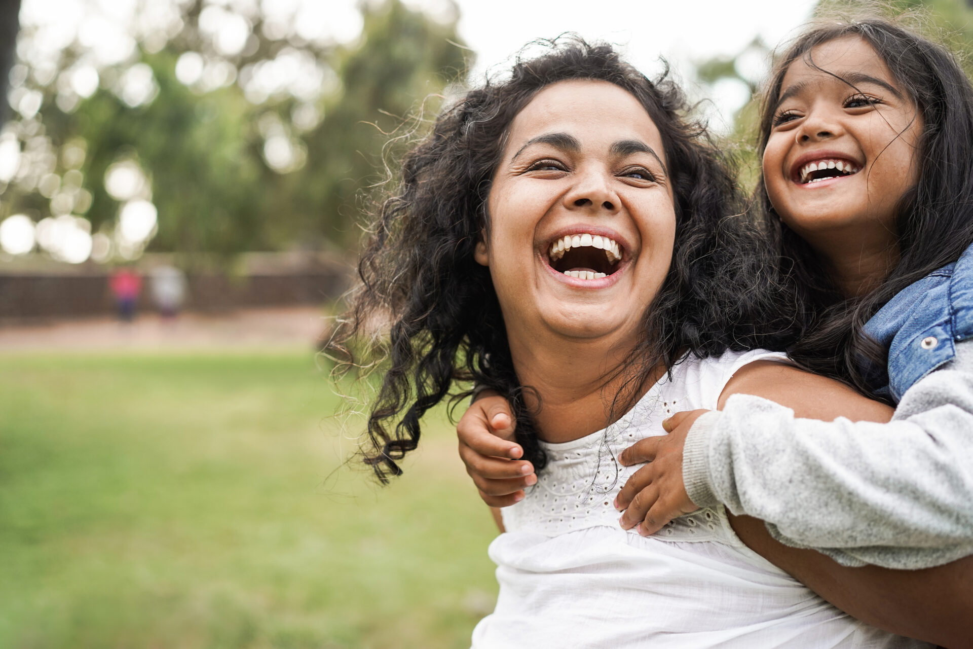 A woman and a young girl with long dark hair laugh joyfully outdoors, with the girl hugging the woman from behind. They are surrounded by greenery, and the background is blurred.