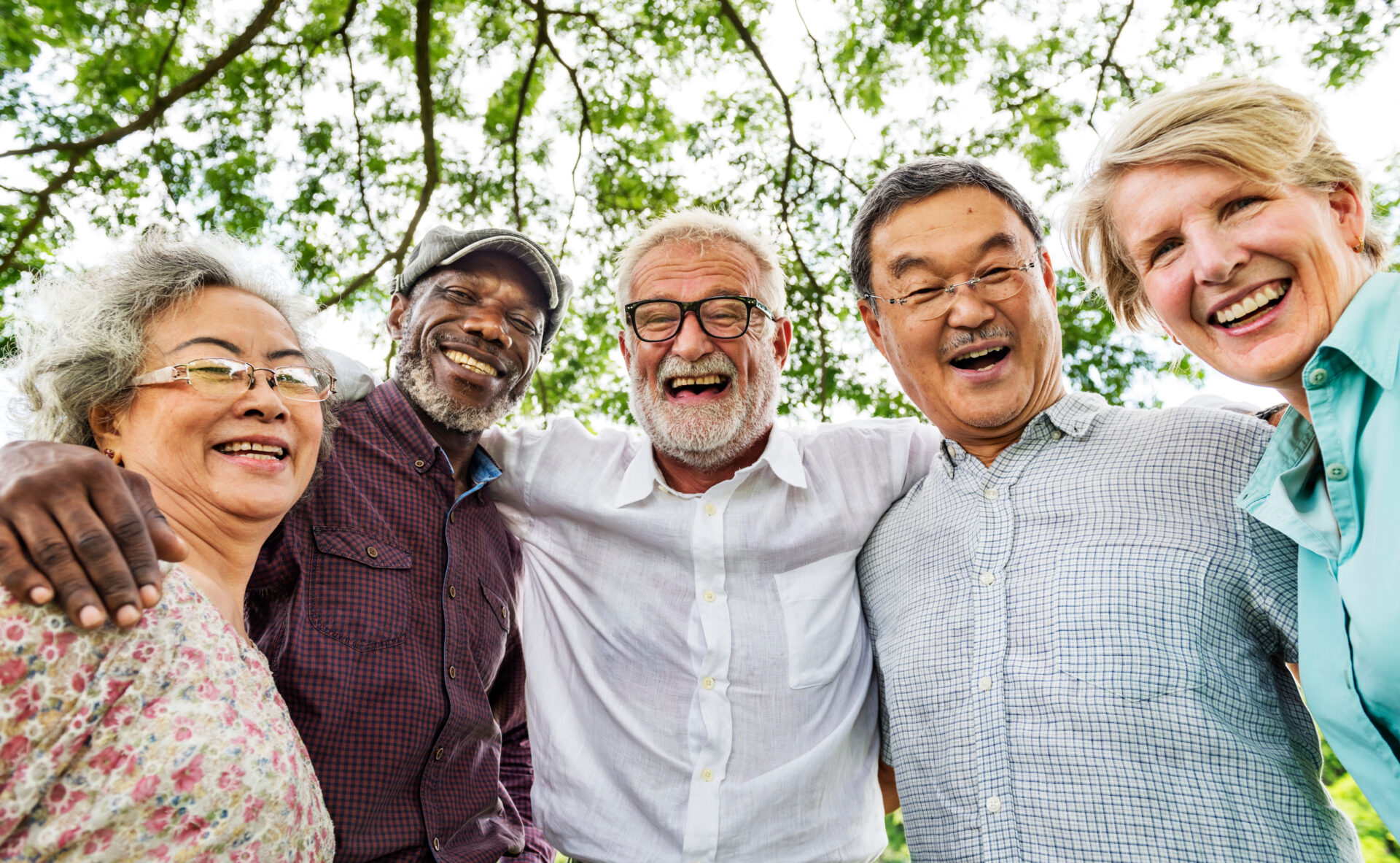 Five older adults stand close together outdoors, smiling and laughing under leafy green trees, with their arms around each other, showing friendship and joy.