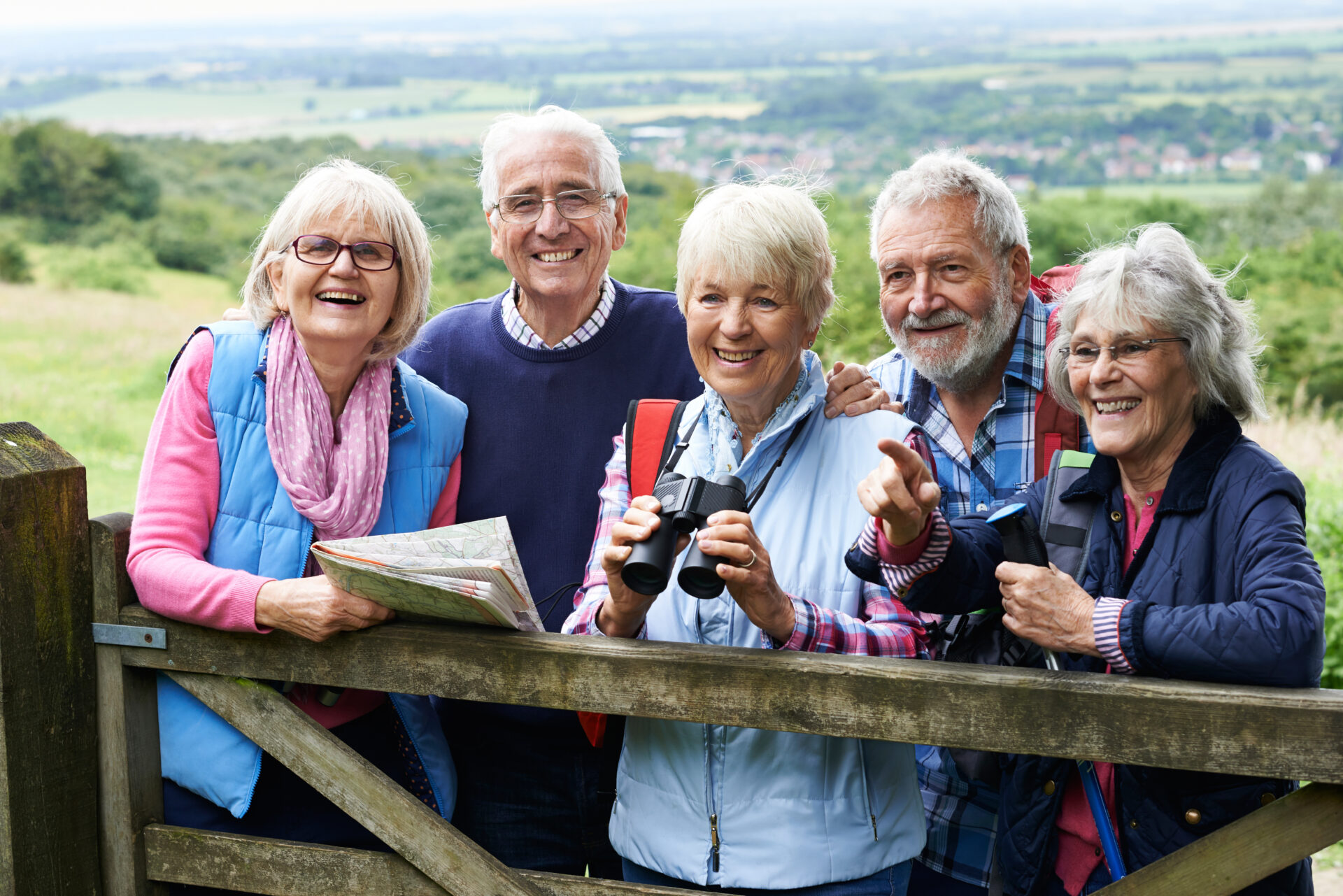 Five smiling older adults stand by a wooden gate in the countryside, with one holding a map and another holding binoculars, enjoying a scenic outdoor day together. Fields and a village are visible in the background.