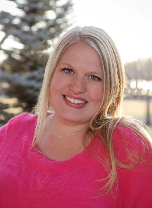A woman with long blonde hair smiles at the camera, wearing a bright pink top and a delicate necklace. She is outdoors with blurred trees and sunlight in the background.