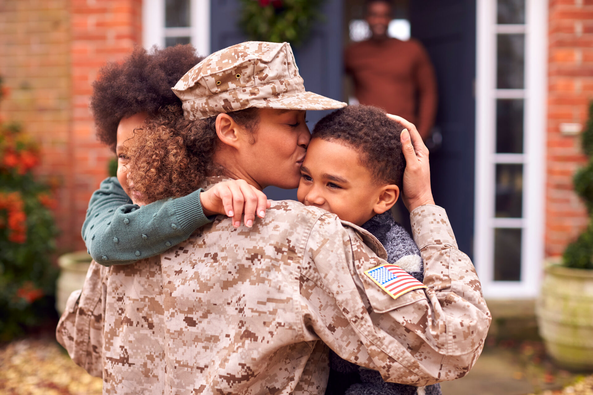 A person in military uniform hugs and kisses two smiling children outside a house, with another adult watching from the doorway in the background.