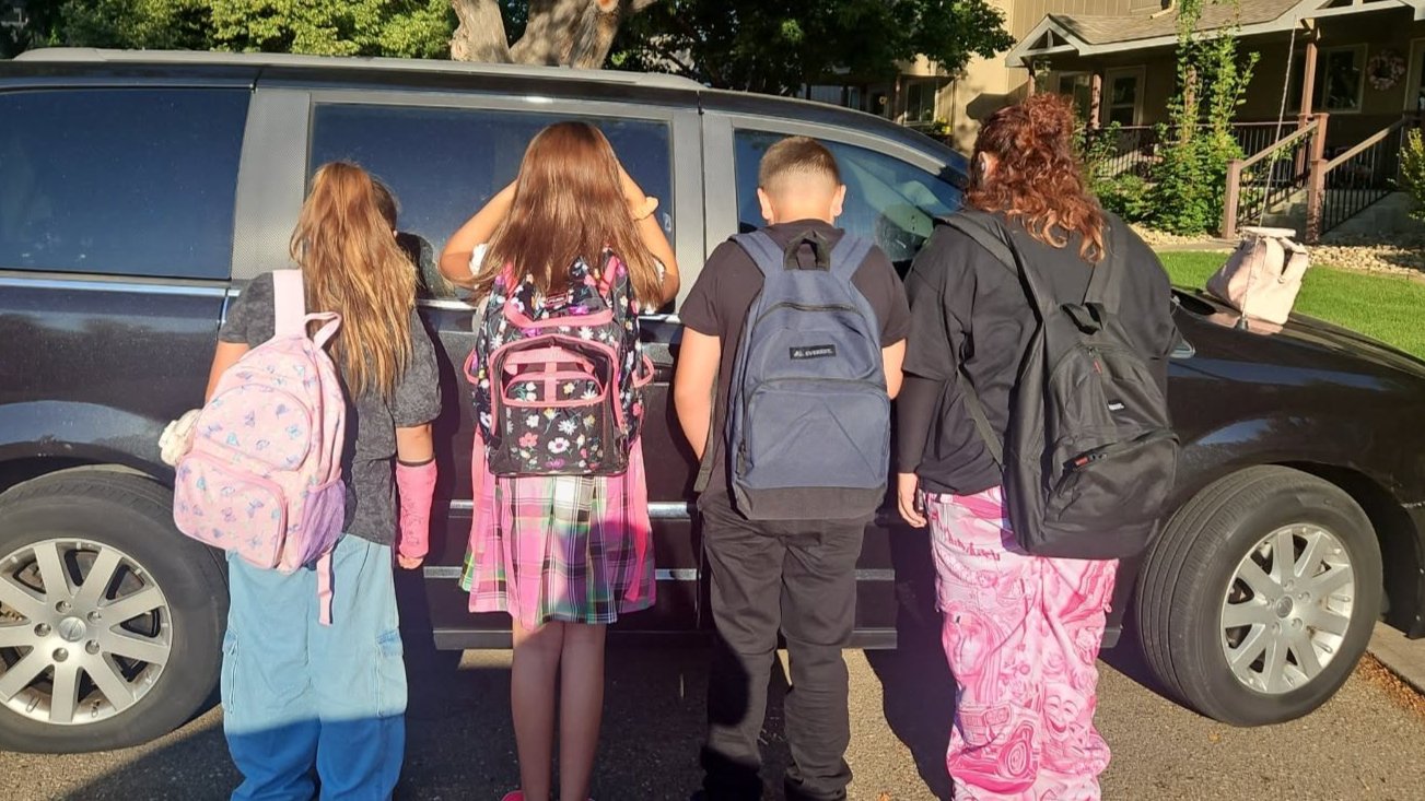 Four children with backpacks stand facing away from the camera toward a black van, dressed in pajamas and colorful outfits, in a suburban neighborhood on a sunny day.