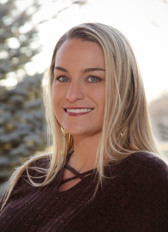 A woman with long blonde hair and blue eyes smiles at the camera, wearing a dark textured top. The background shows blurred trees and soft natural light.