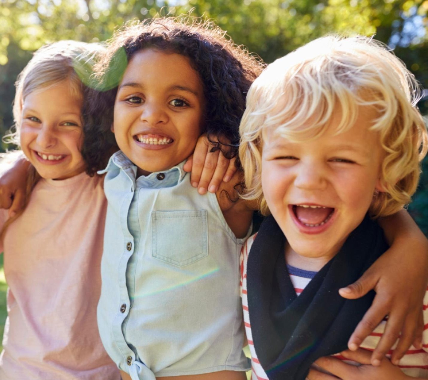 Three young children stand close together outdoors, smiling and laughing with their arms around each other’s shoulders. They appear happy and are bathed in natural sunlight with a green, leafy background.