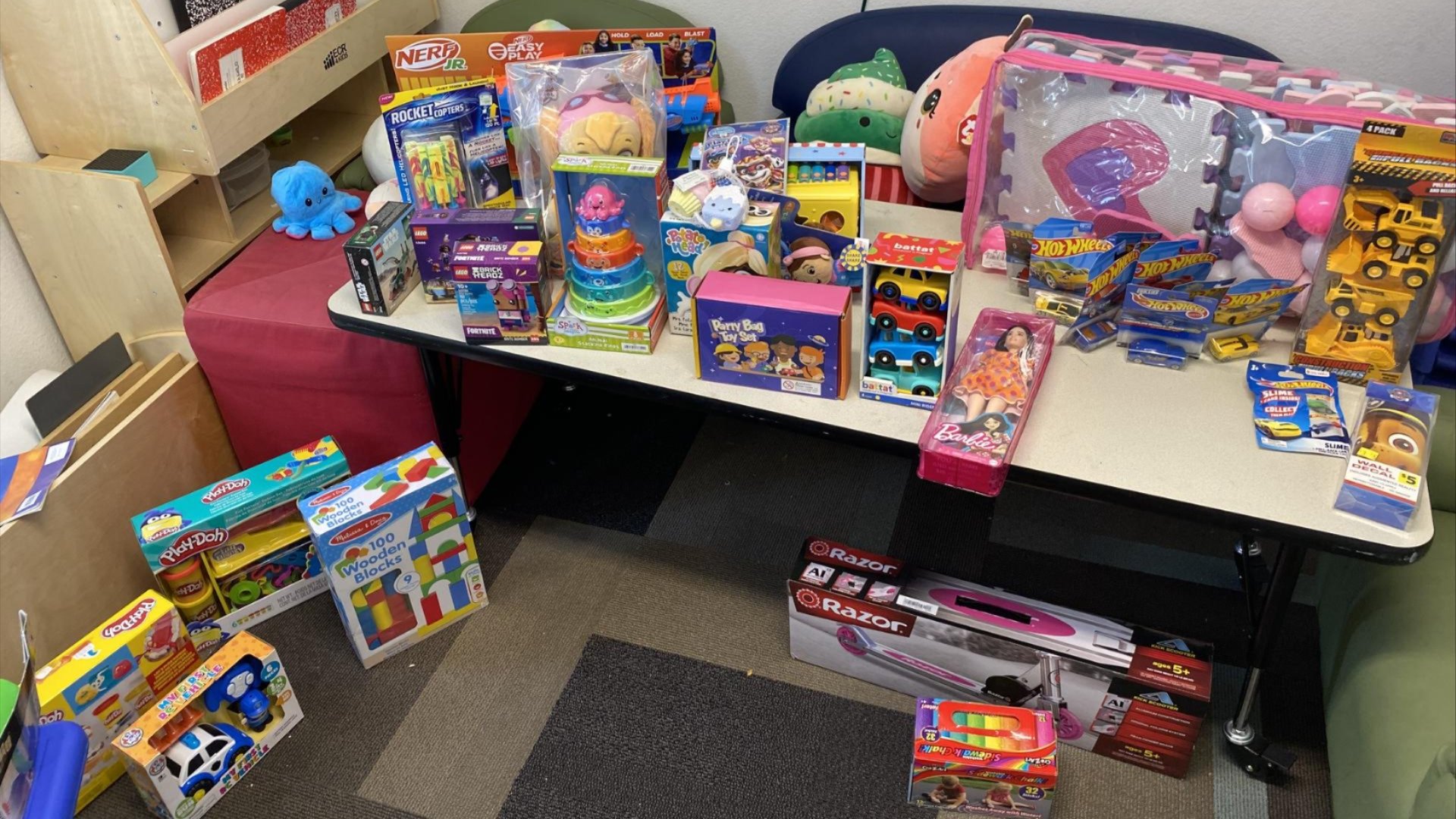 A table and floor filled with a variety of boxed and packaged toys, including Nerf blasters, Hot Wheels cars, Barbie dolls, Play-Doh, and plush animals in a classroom or playroom setting.