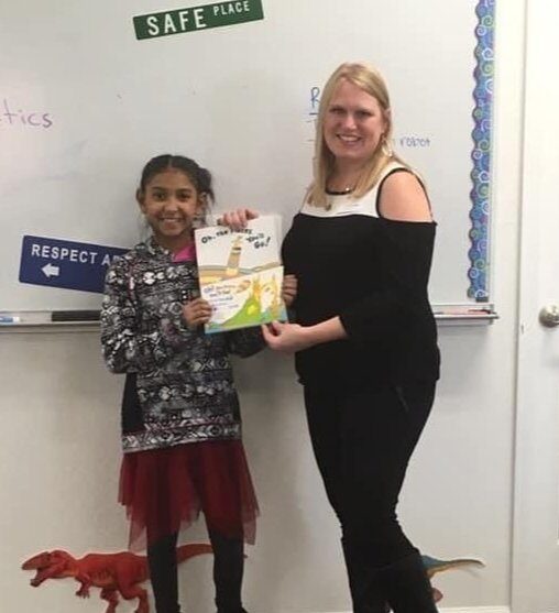 A young girl and a woman stand smiling in a classroom, holding a book titled "Go, Go, Go!" There are signs reading "SAFE PLACE" and "RESPECT" on the whiteboard behind them. A toy dinosaur is on the floor.