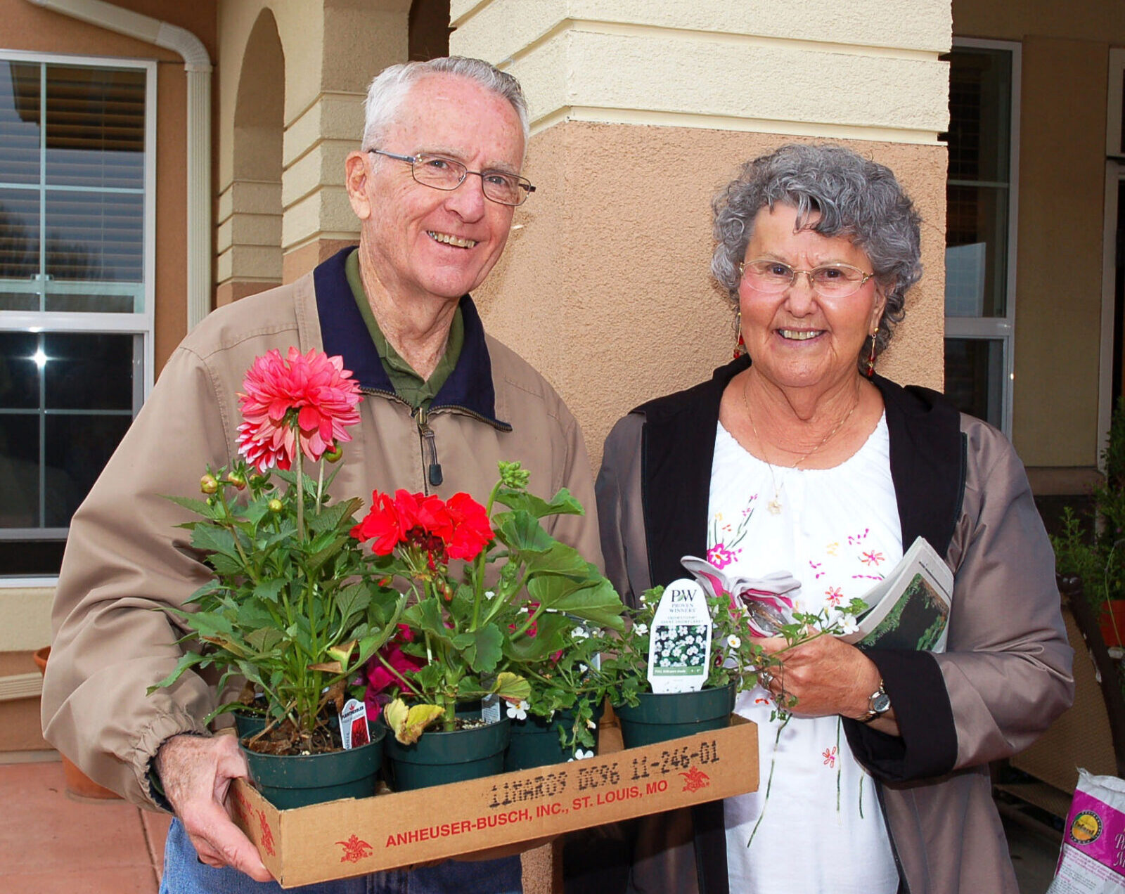 An older man and woman stand outside a house, smiling. The man holds a cardboard tray with potted flowers and gardening supplies. The woman holds small flowers and a booklet.