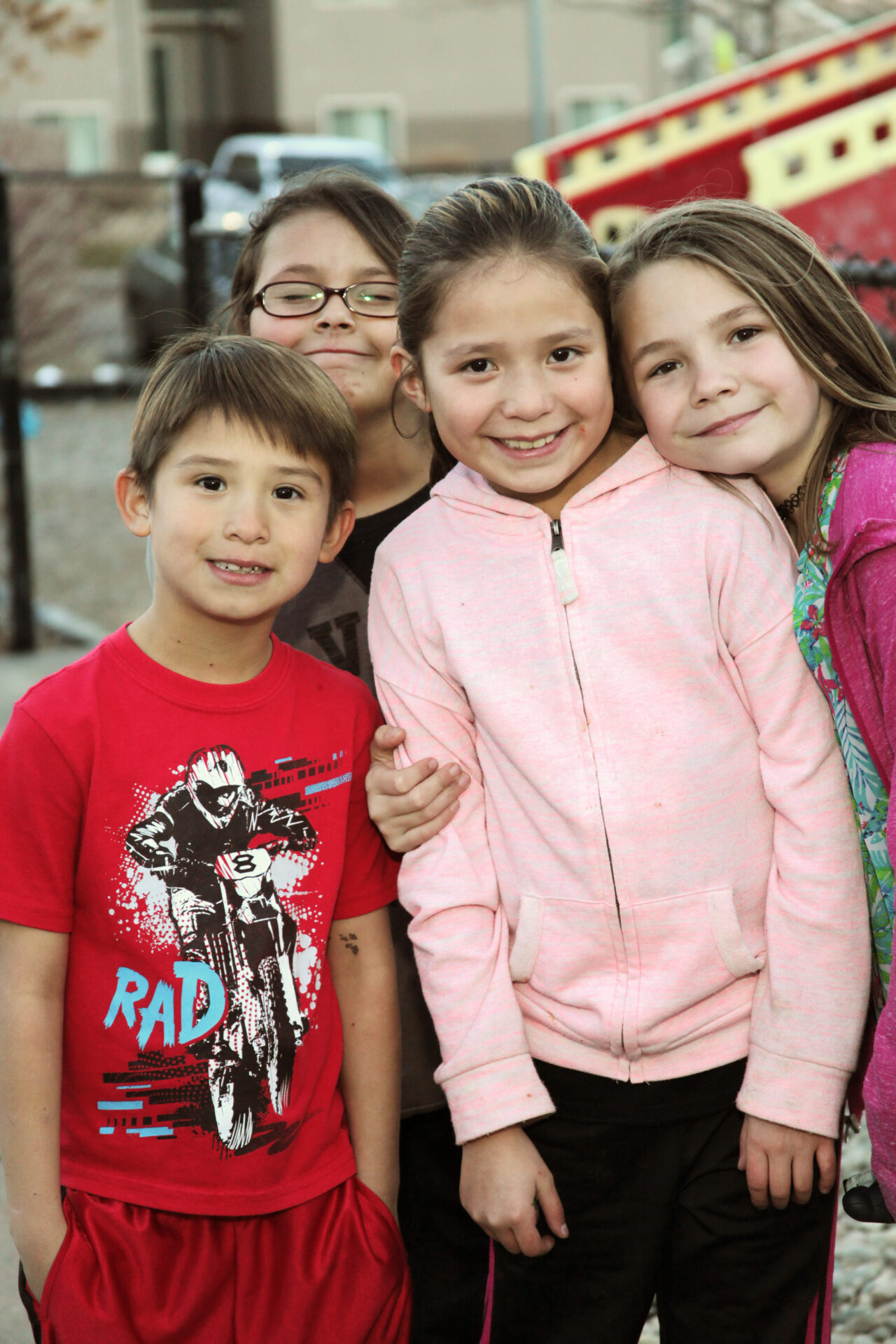 Four children stand close together outdoors, smiling at the camera. Two girls wear pink jackets, another girl has glasses, and the boy at the front wears a red shirt with a motorcycle graphic. A playground is visible in the background.