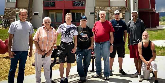 A group of eight people, mostly men, stand together outside a modern three-story apartment building on a cloudy day, posing and smiling for the camera.