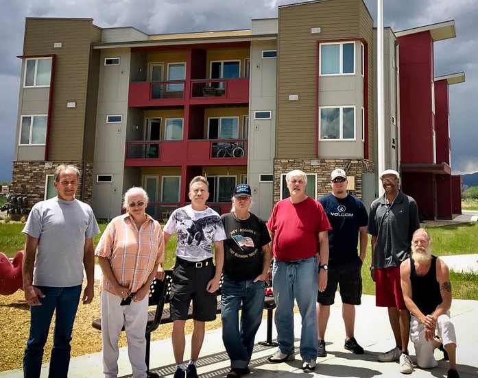 A group of eight people, mostly men, stand together outside a modern three-story apartment building on a cloudy day, posing and smiling for the camera.