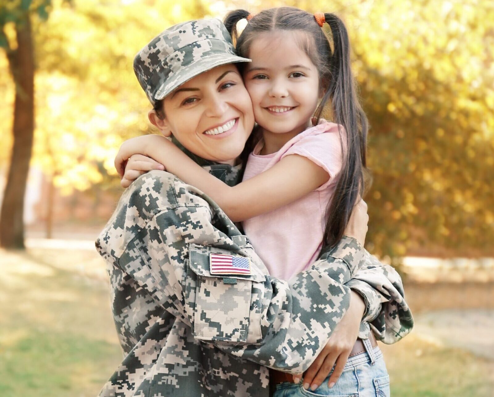 A woman in military uniform hugs a smiling young girl outdoors. The woman wears camouflage and a cap, and the girl has long brown hair in pigtails. They both appear happy, with trees and sunlight in the background.