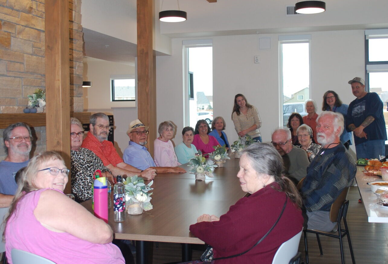 A group of people, mostly seniors, sit around a long table in a bright, modern room with wooden beams. They are smiling and appear to be gathered for a social event or meal.