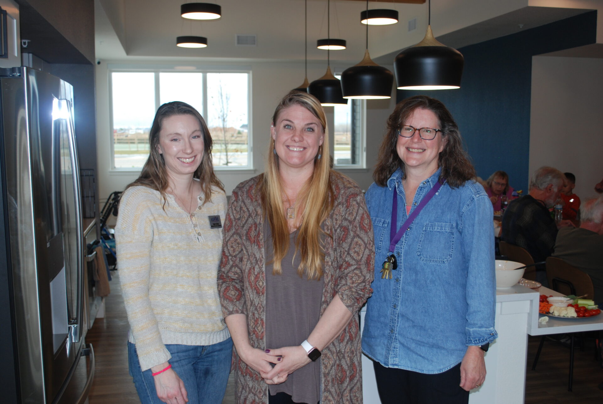 Three women smiling and standing together in a modern kitchen with pendant lights. People are seated and talking in the background. The atmosphere appears casual and friendly.