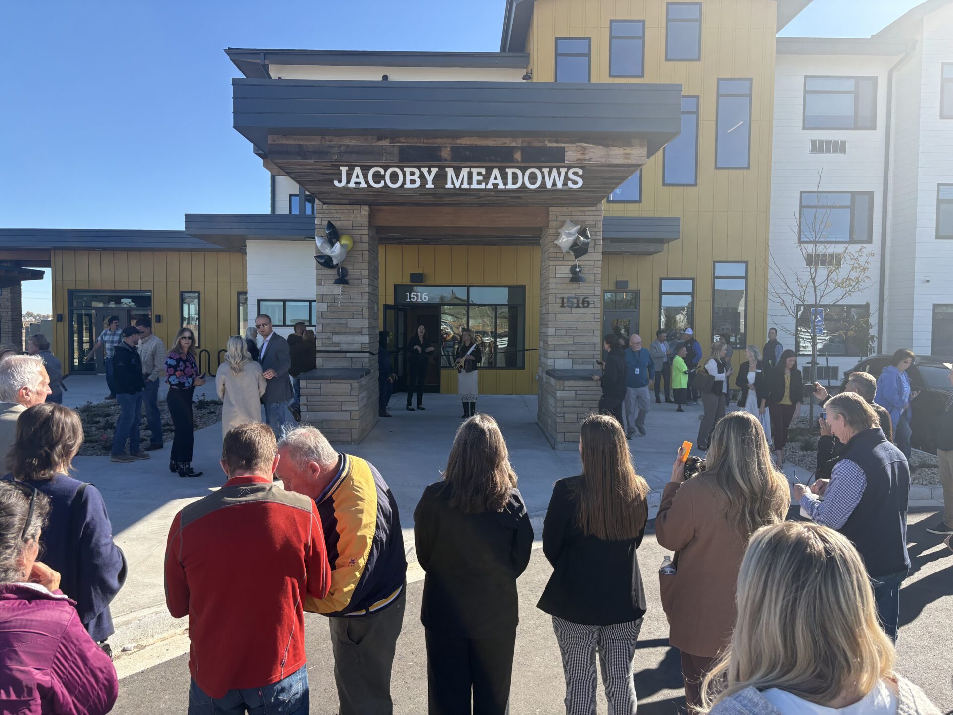 A group of people stands outside the entrance of a modern building with a sign reading "Jacoby Meadows" above the door, attending what appears to be an outdoor event or ceremony on a sunny day.