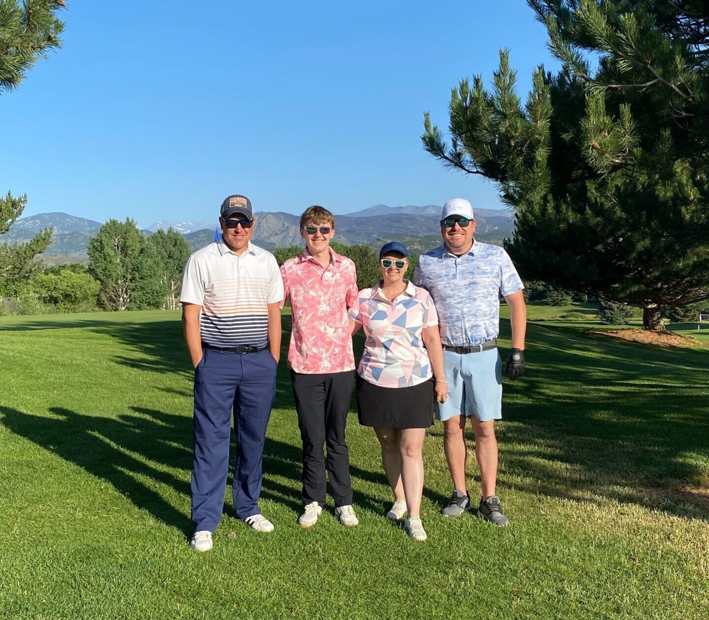 Four people stand side by side on a sunny golf course, each wearing golf attire. Trees and mountains are visible in the background under a clear blue sky.