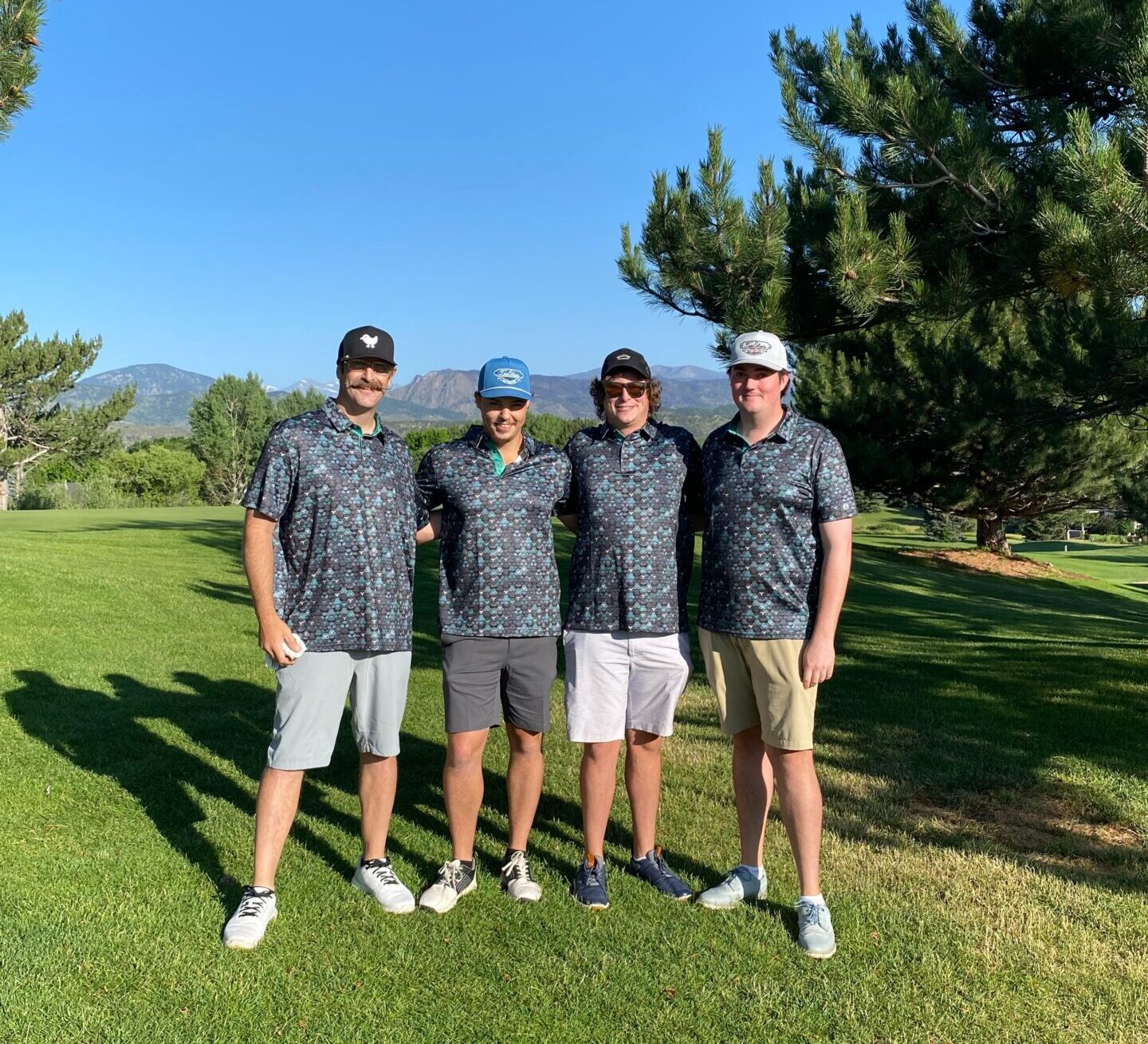 Four men stand together on a sunny golf course, all wearing matching patterned shirts and hats. Trees and blue sky are in the background, with distant mountains visible.
