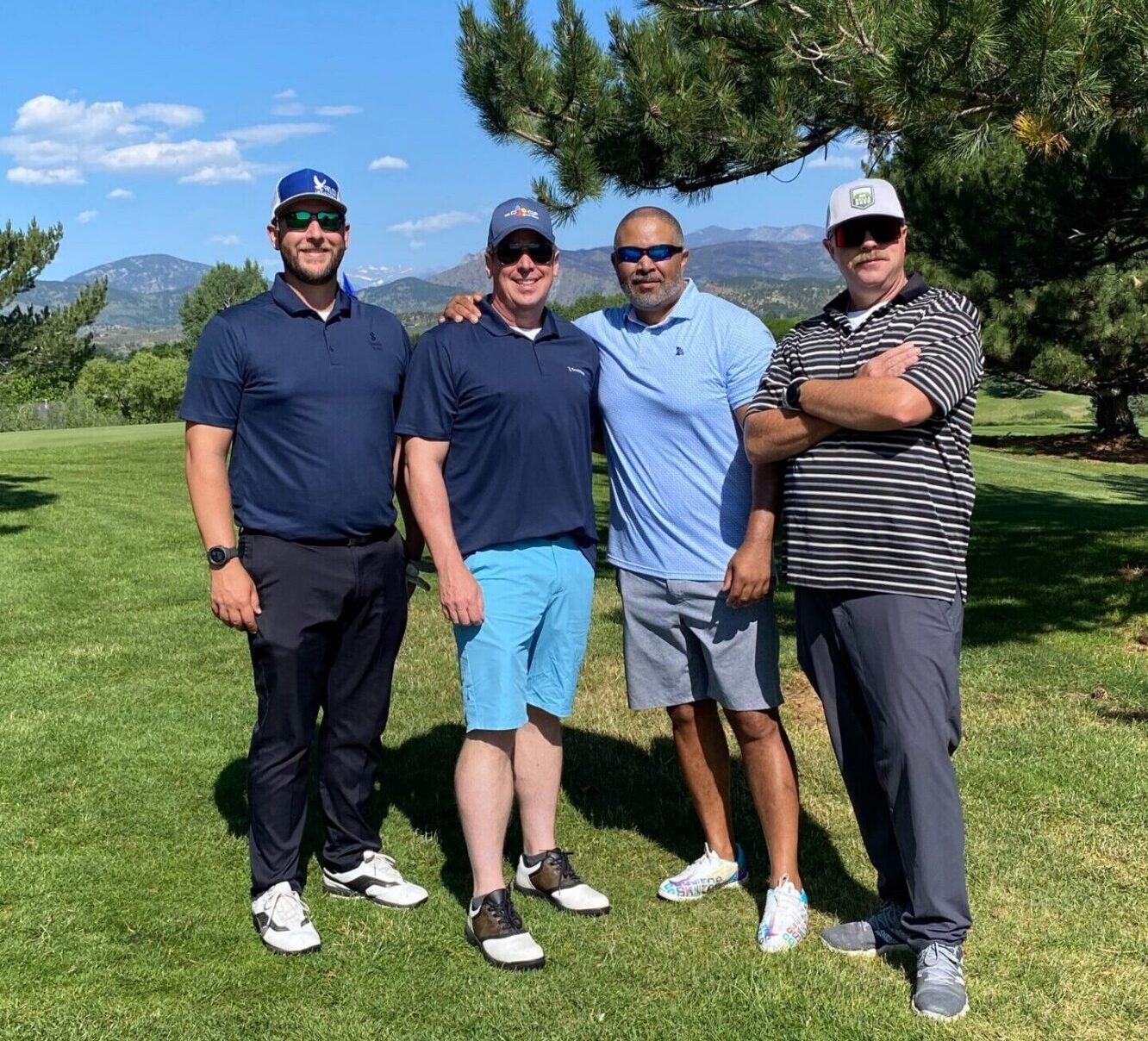 Four men stand together on a sunny golf course with trees and mountains in the background, all wearing casual golf attire, including polo shirts, shorts or pants, and sunglasses or hats.