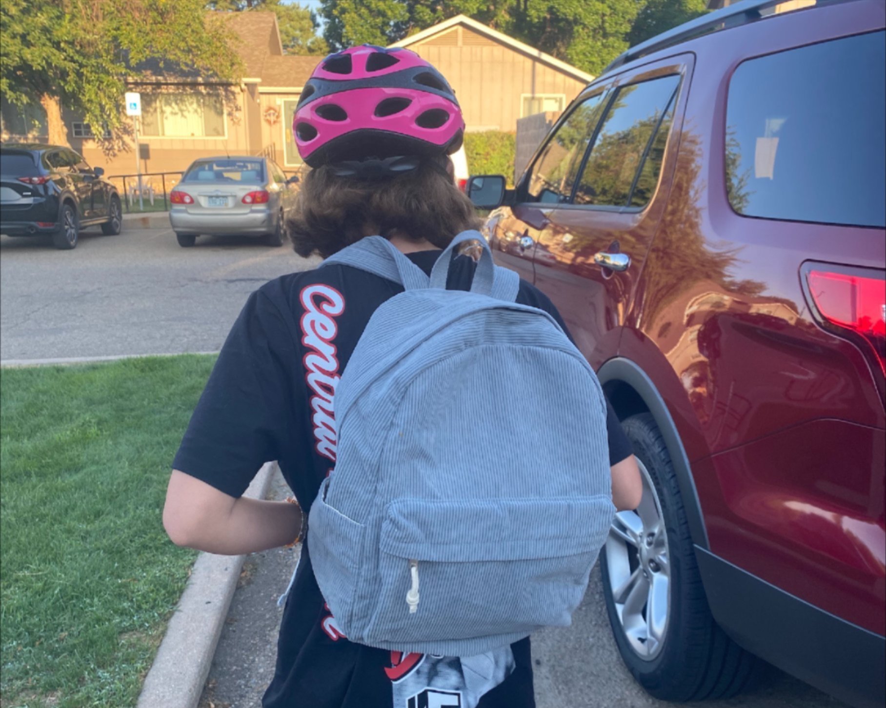 A child wearing a pink bike helmet and a gray backpack walks on a sidewalk next to a red SUV, with houses and other parked cars visible in the background.