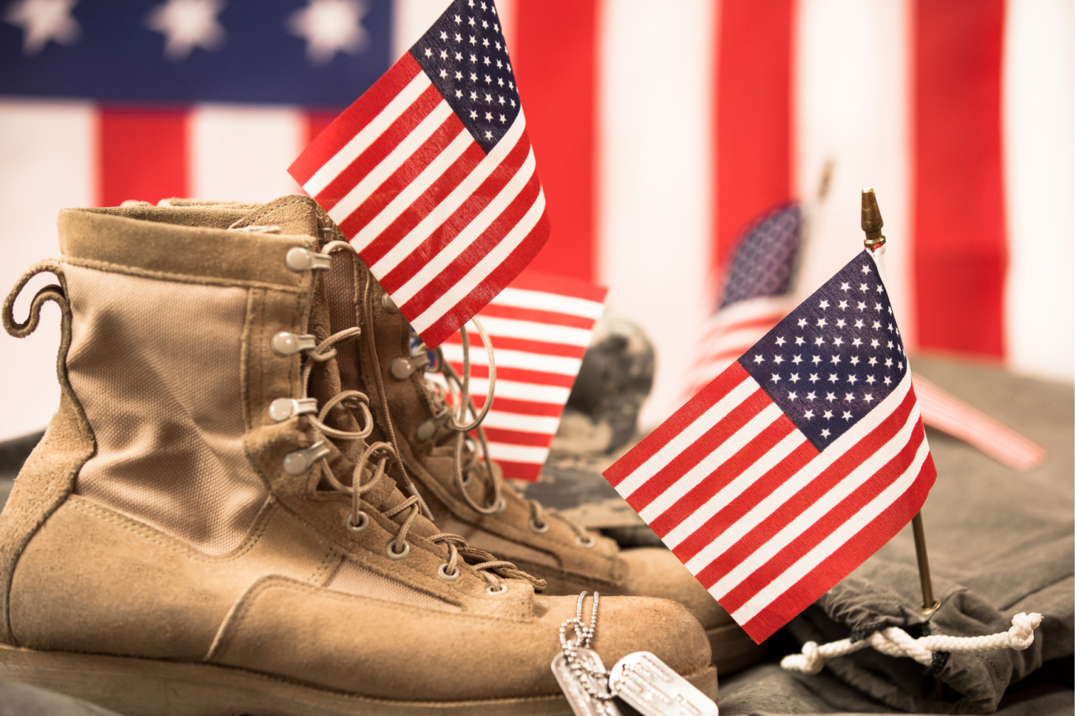 A pair of tan military boots with American flags and dog tags placed on top, set against a blurred background of the U.S. flag.
