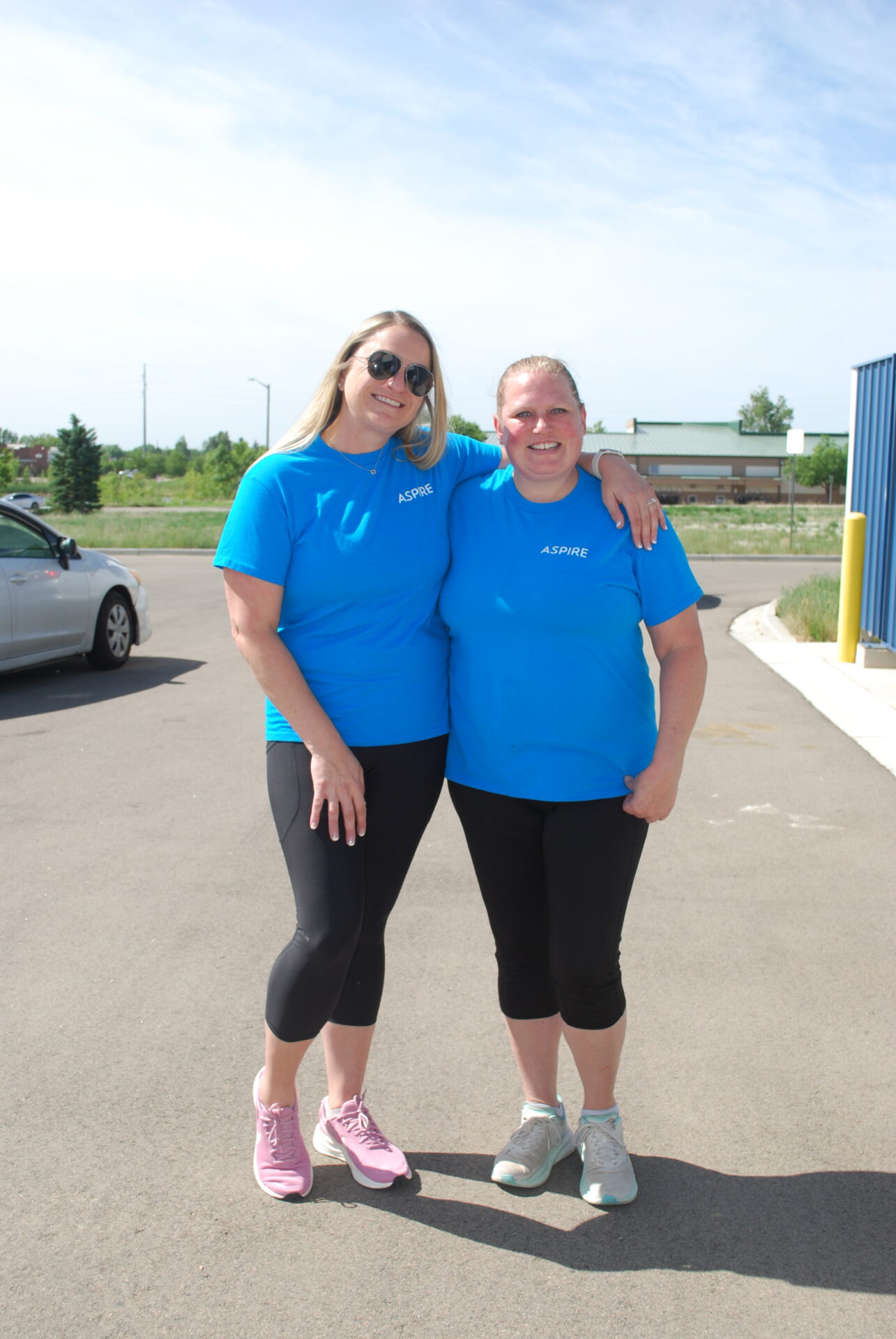 Two women smile outdoors, both wearing matching bright blue "Aspire" T-shirts, black leggings, and sneakers. One woman has her arm around the other's shoulders. A car and building are visible in the background.