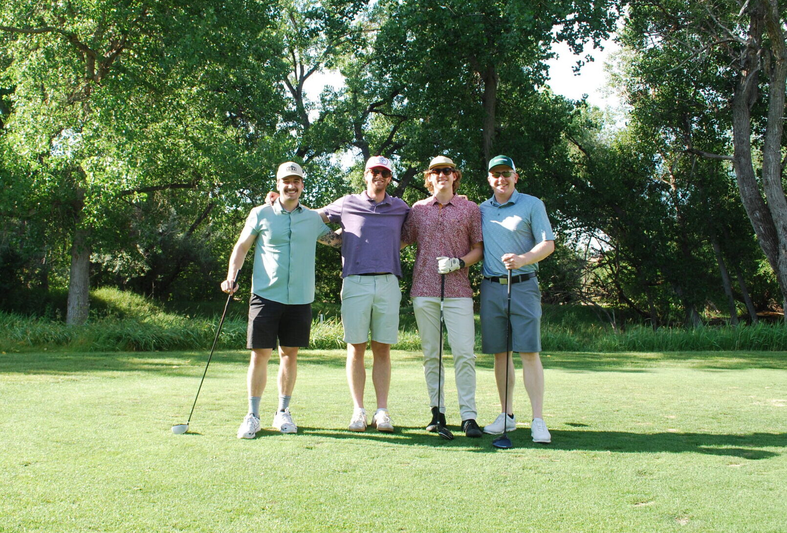 Four people stand together on a golf course, smiling at the camera. They wear casual golf attire and hats, and one holds a golf club. Trees and grass fill the sunny background.
