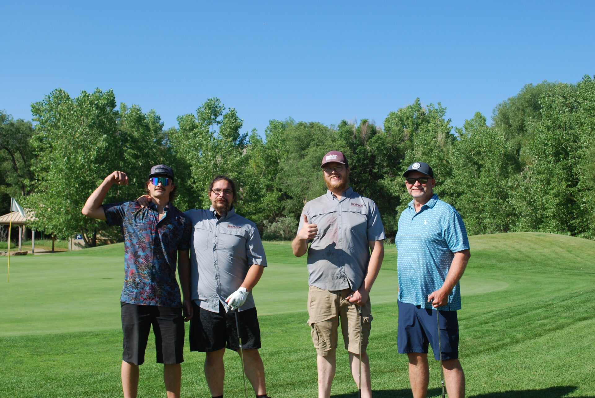 Four men stand on a golf course green under a clear blue sky, holding golf clubs and making hand gestures, with trees and a small structure in the background.
