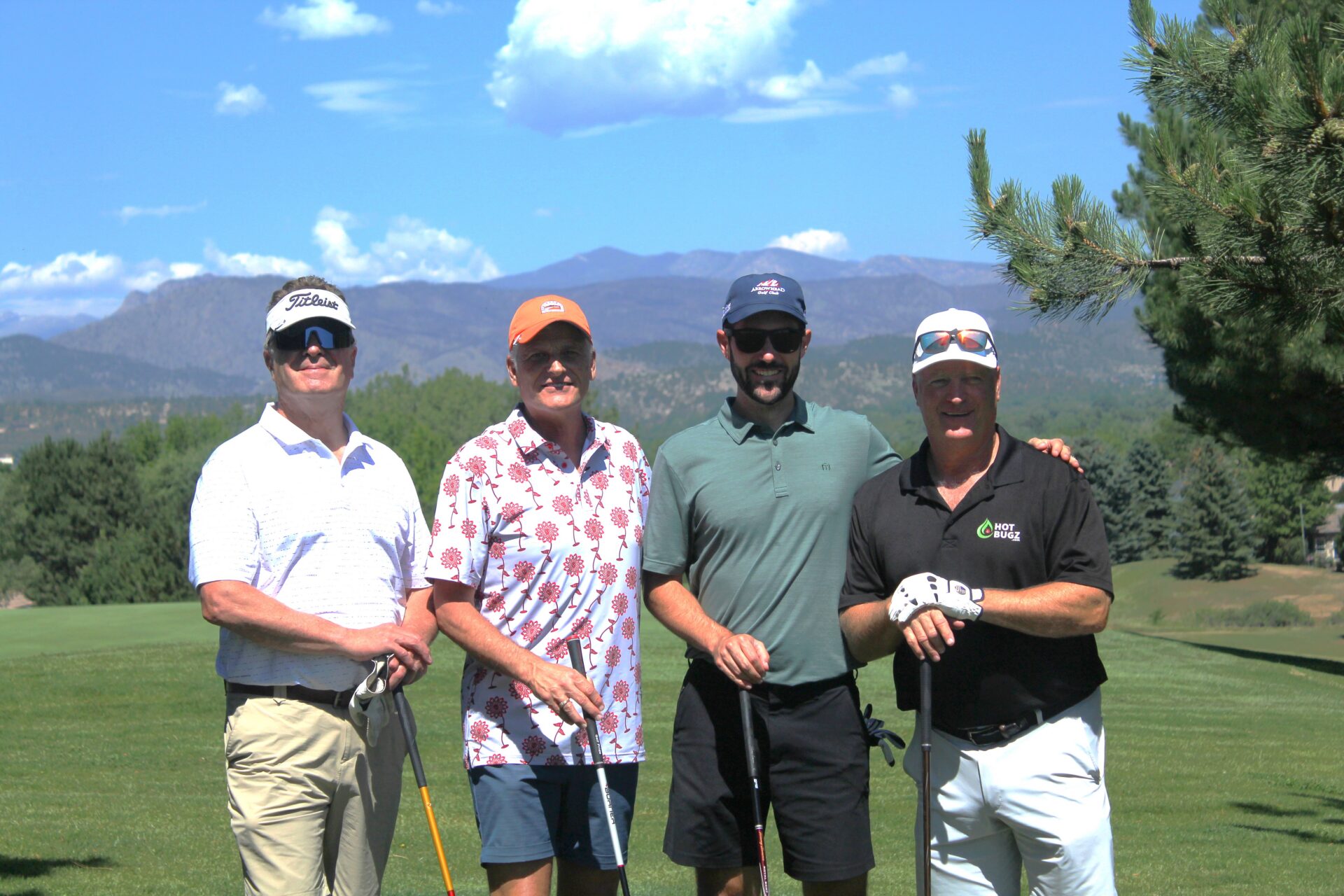 Four men pose together on a sunny golf course with mountains in the background. They are dressed in golf attire and holding clubs, smiling for the photo.
