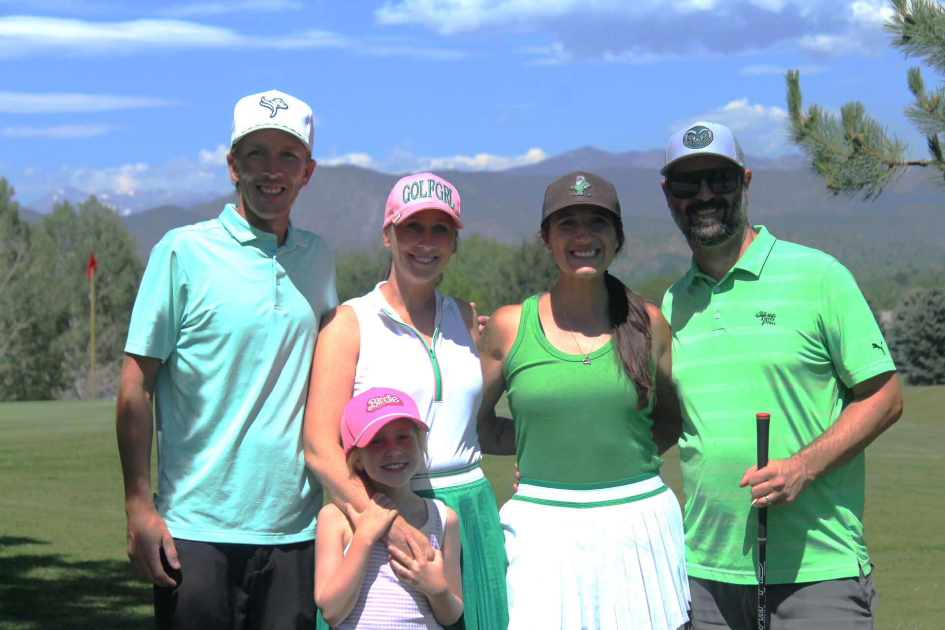 Five people, including a young girl, stand and smile on a sunny golf course with green grass, trees, and mountains in the background. Most wear green and white golf attire and caps.