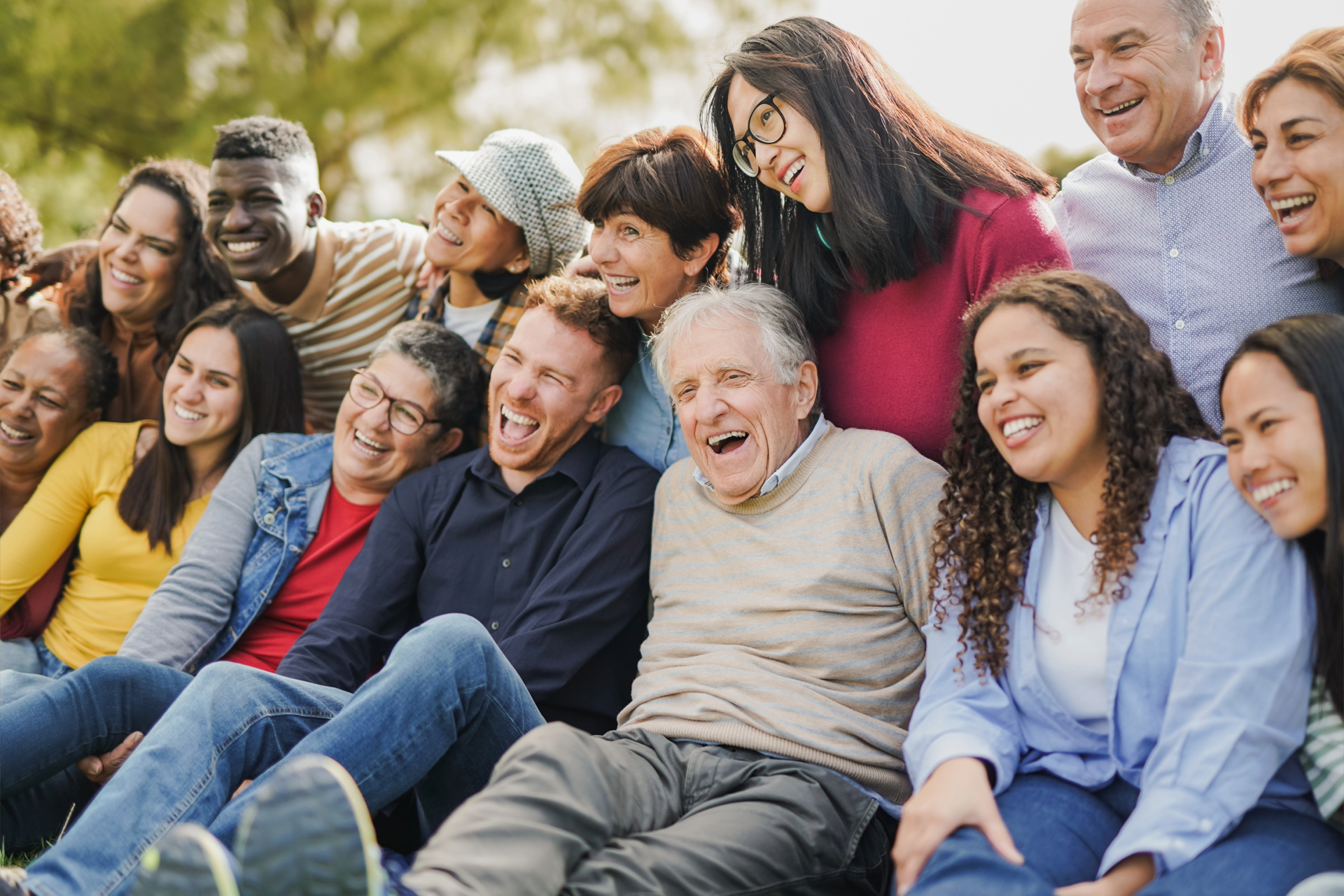 A diverse group of people of various ages sit closely together outdoors, smiling and laughing, appearing joyful and connected on a sunny day.