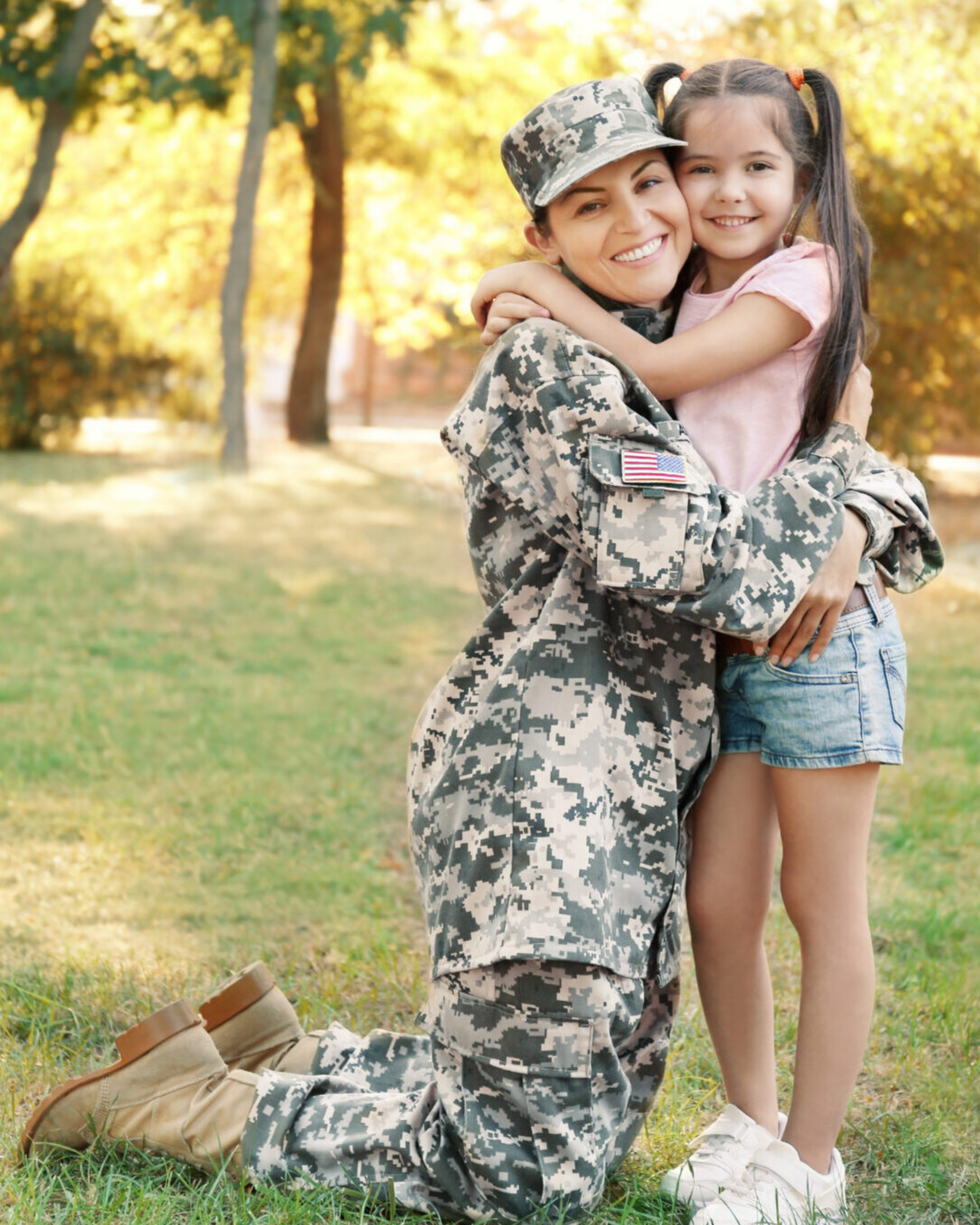 A smiling woman in military uniform kneels on grass, hugging a young girl who stands beside her. They are outdoors in a sunlit park with green trees in the background.