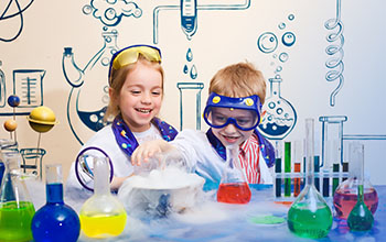 Two children wearing safety goggles and lab coats smile while conducting a science experiment with colorful liquids and fog in beakers, with a chemistry-themed background.