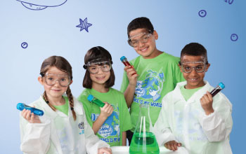 Four children wearing safety goggles and green science-themed shirts hold colorful test tubes, standing around a table with a green liquid in a beaker. Blue science doodles are sketched on the light blue background.