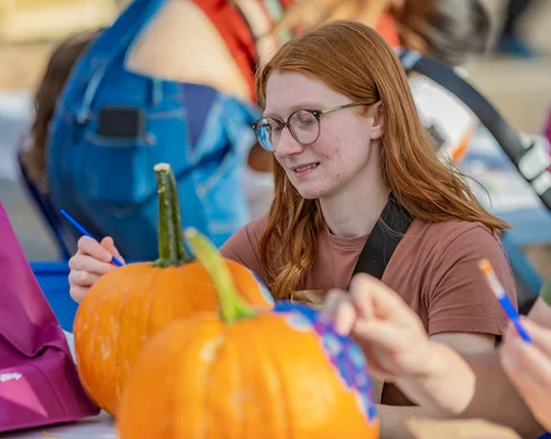 A person with long red hair and glasses paints designs on pumpkins at a table during an outdoor event. Other people and pumpkins are visible in the background.